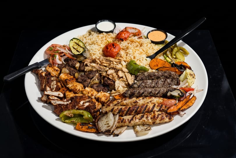 A large platter of grilled meats, rice, roasted vegetables, and sauces on a white plate.