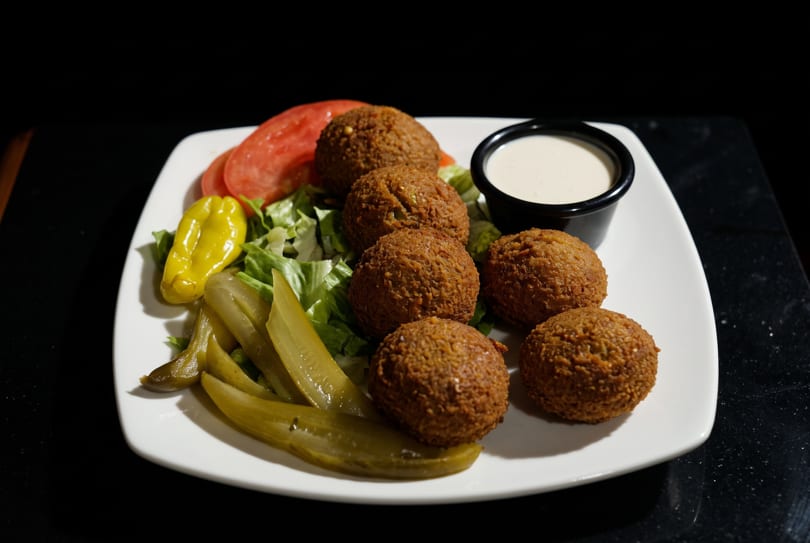 A plate of falafel with lettuce, tomato, pickles, a yellow pepper, and a side of white sauce.