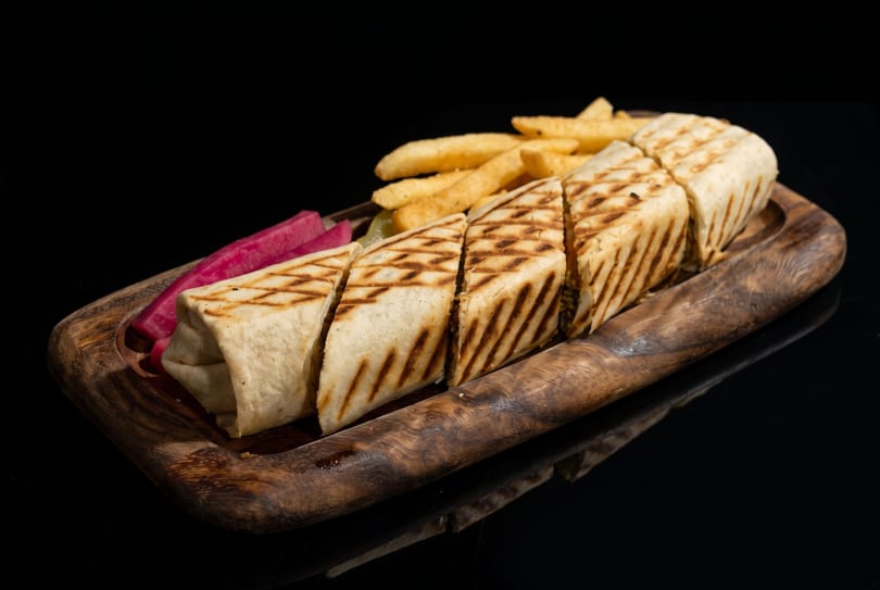Grilled chicken shawarma wrap, cut into pieces, with french fries and pickled turnips on a wooden tray.