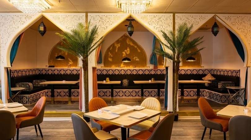 Restaurant interior with ornate archways, palm trees, and tables set with white plates.